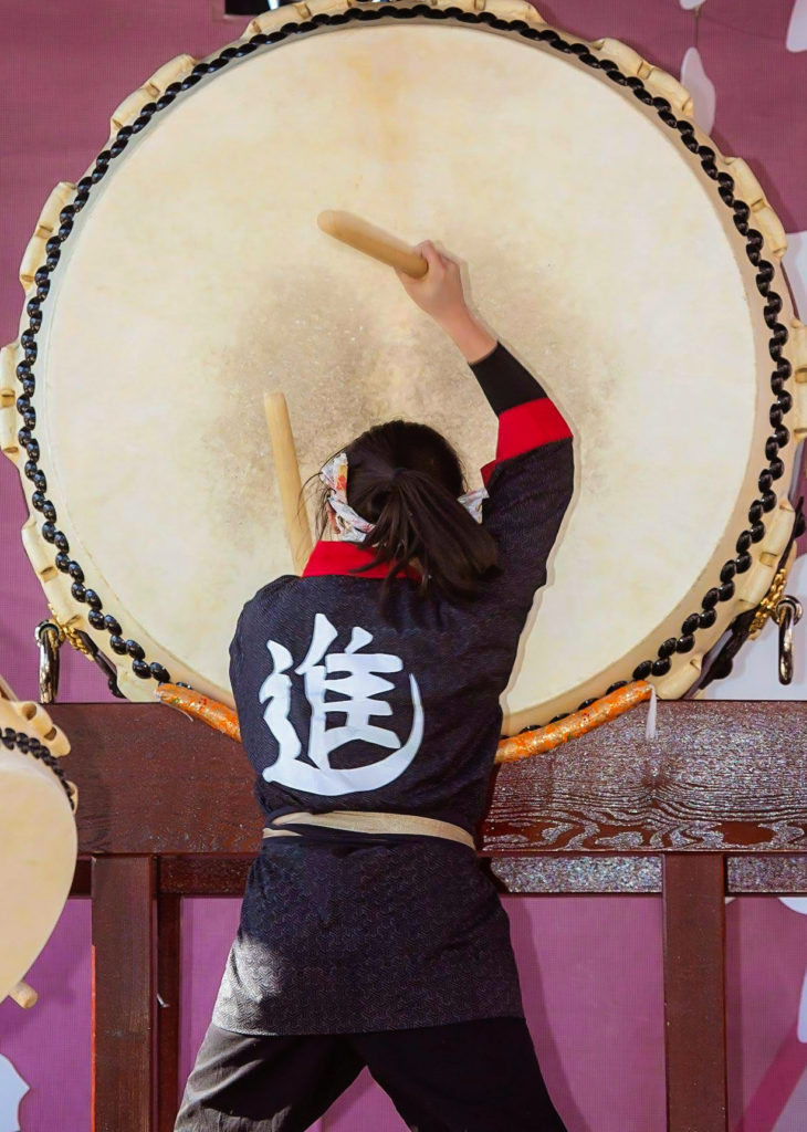 A woman dressed in traditional Japanese clothing plays a large Japanese drum, or odaiko