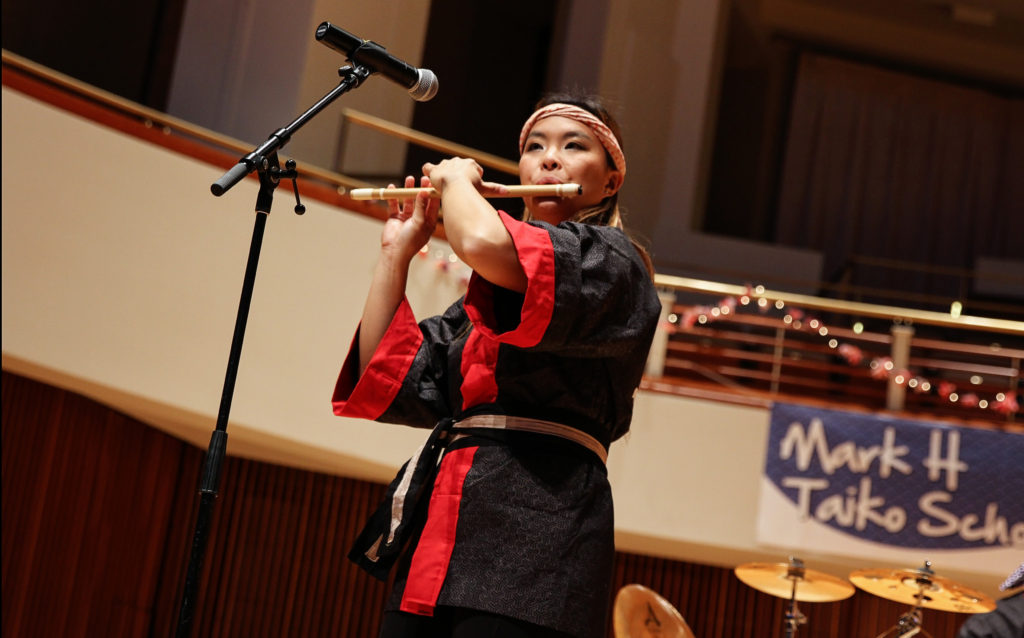 A woman dressed in traditional Japanese clothing plays a bamboo flute, or shinobue